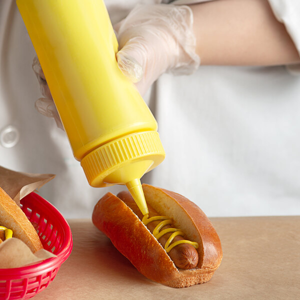 A person using a Vollrath yellow wide mouth squeeze bottle to put mustard on a hot dog.