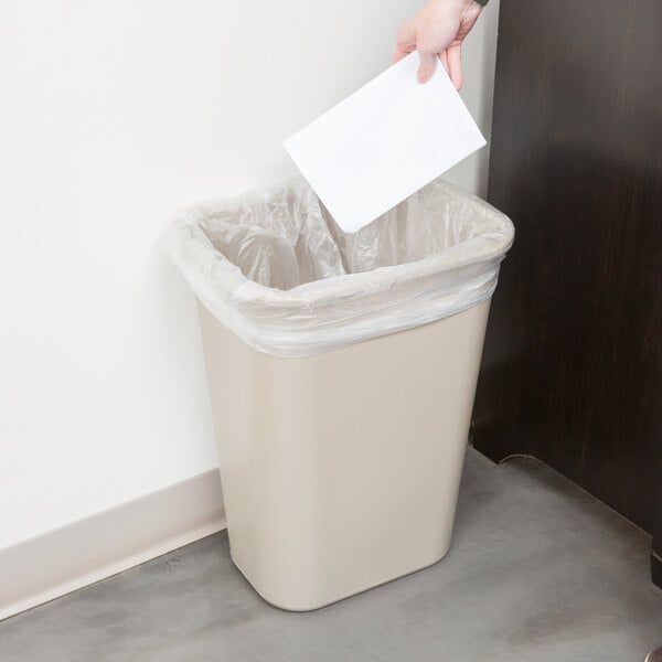 A hand throws a piece of paper into a Rubbermaid beige rectangular trash can.