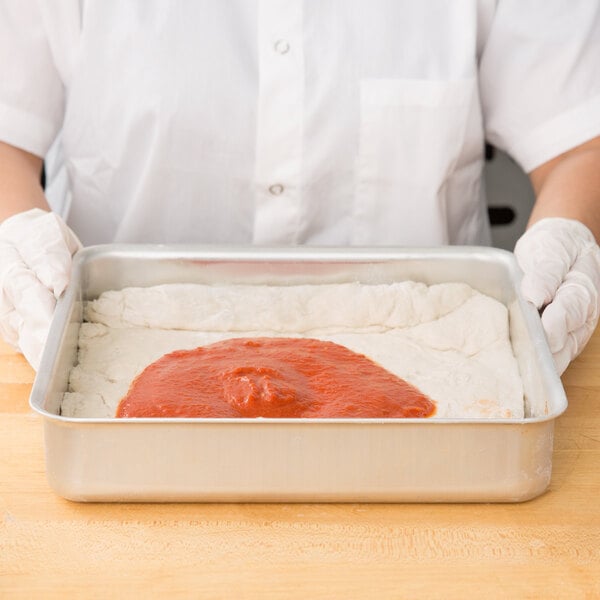 A person in white gloves holding a pan of pizza dough with red sauce in an American Metalcraft aluminum cake pan.