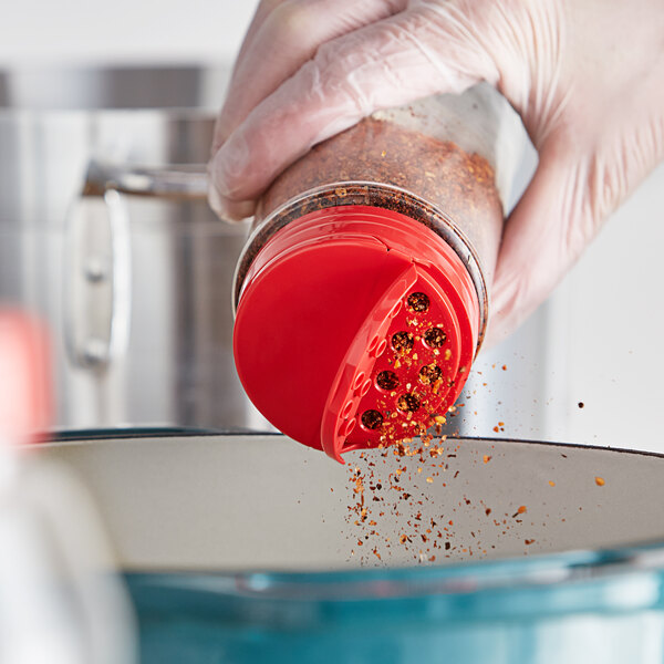 A person using a red dual-flapper spice lid to pour red flakes into a bowl.