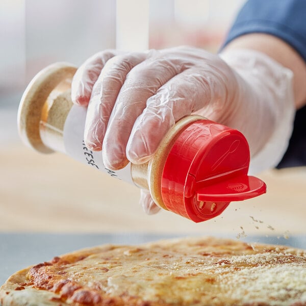 A hand in gloves using a 53/485 round red plastic spice container to sprinkle seasoning on a pizza.