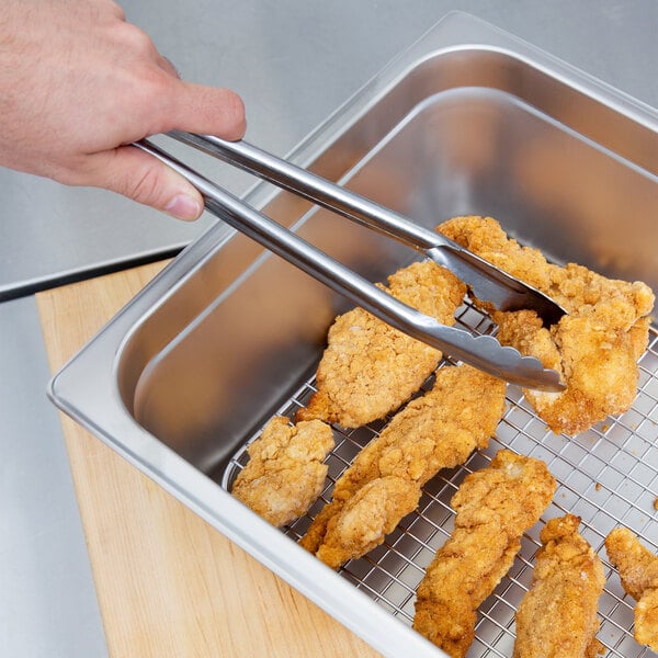 A person using tongs to hold fried chicken over a stainless steel pan grate.