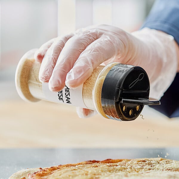 A person in a white glove holding a 7 oz. round plastic spice container with a shaker lid over a pizza.