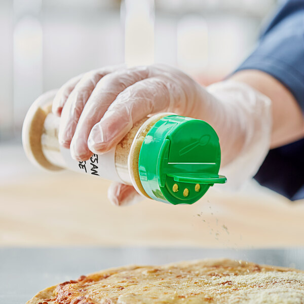 A hand in a glove pouring seasoning from a 53/485 round plastic spice container with a green dual flapper lid onto a pizza.