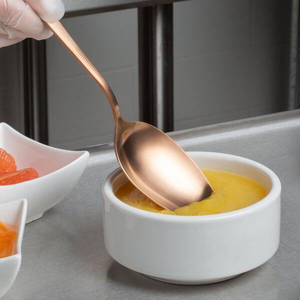 A person's hand holding a Mercer Culinary rose gold plating spoon over a bowl of food.