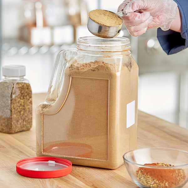 A person pouring brown powder into a rectangular plastic spice container with a red lid.