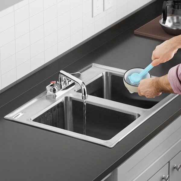 A person is washing a bowl at a double-basin stainless steel kitchen sink with water running from the faucet.