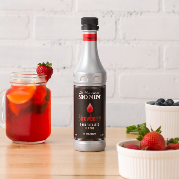 A close-up of a Monin Strawberry Concentrated Flavor bottle next to a bowl of strawberries.
