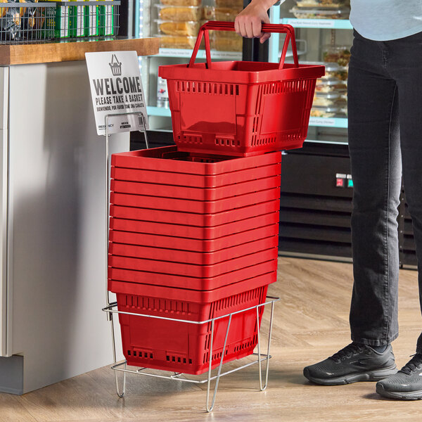 A stack of red plastic shopping baskets with handles is placed on a metal stand next to a sign inviting customers to take a basket.