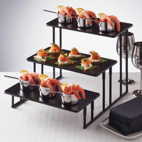 An American Metalcraft 3-tier black rectangular riser stand holding food on a table.