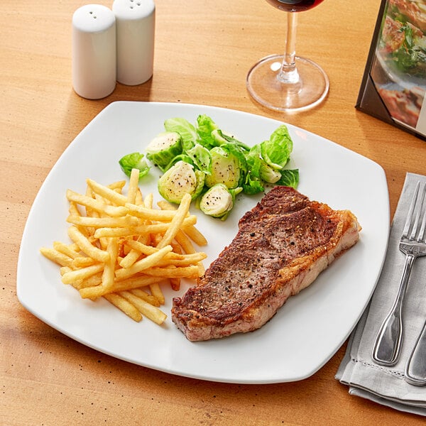 A close-up of an Acopa Bright White square porcelain coupe plate with steak, fries, and vegetables on it with a glass of wine.
