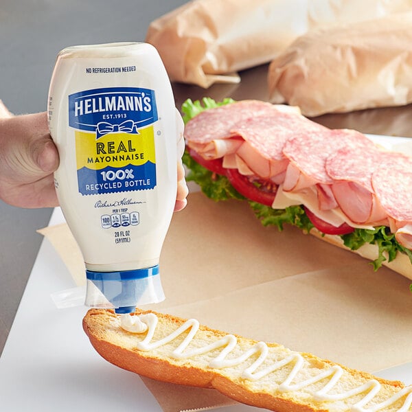 A hand holding a white Hellmann's mayonnaise bottle over a sandwich.