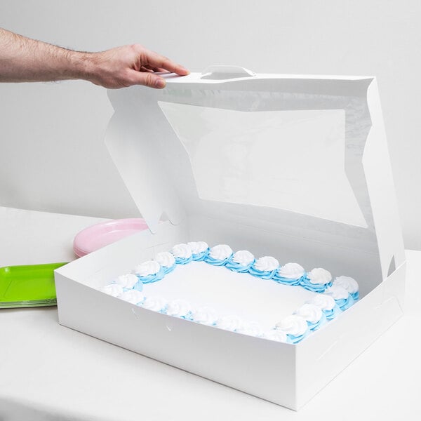 A hand opening a white window cake box to reveal a white and blue cake.