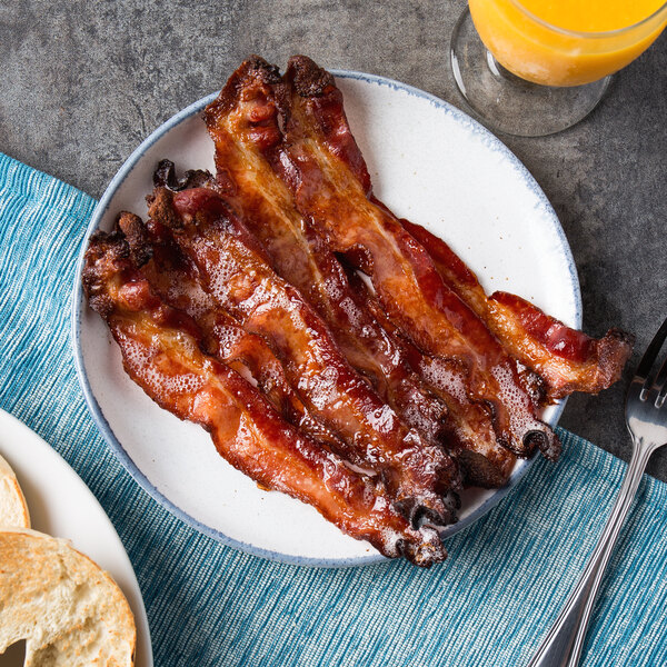 A plate of Kunzler thick apple wood smoked cinnamon sliced bacon with a glass of orange juice on a white table.