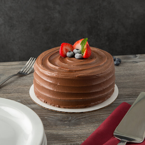 A chocolate cake with strawberries and blueberries on top sits on a corrugated white cake circle on a table in a bakery display.