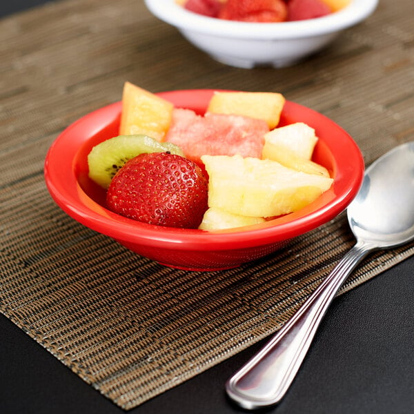A Carlisle red rimmed melamine fruit bowl filled with fruit on a table with a spoon.