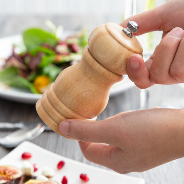 A person holding an Acopa matte natural wooden pepper mill over a plate of food.