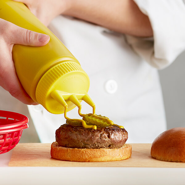 A hand holding a Vollrath Tri Tip Squeeze Bottle with yellow cap pouring mustard onto a burger.
