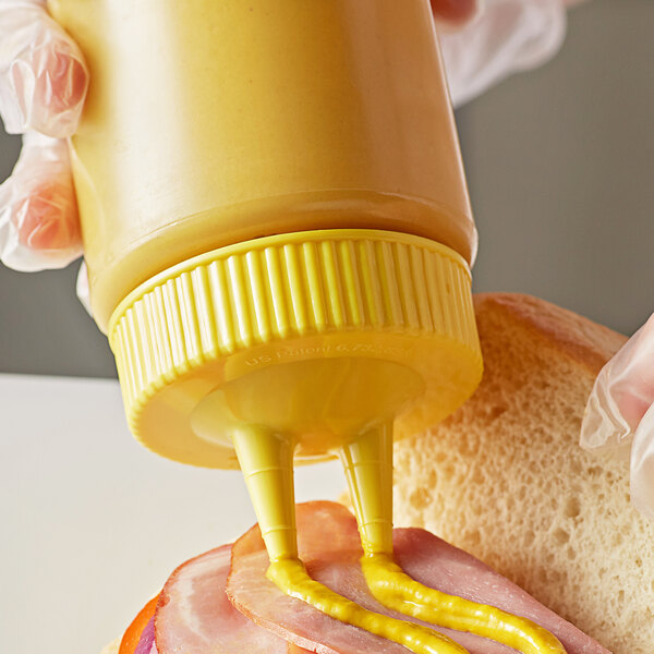 A person using a Vollrath Clear Twin Tip Squeeze Bottle with Yellow Cap to put mustard on a sandwich.