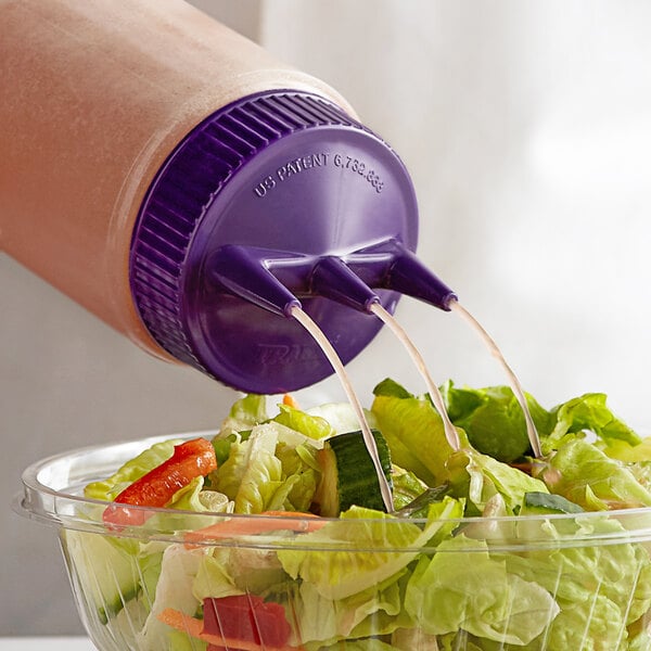 A clear Vollrath Tri Tip wide mouth squeeze bottle with a purple cap pouring dressing onto a salad.