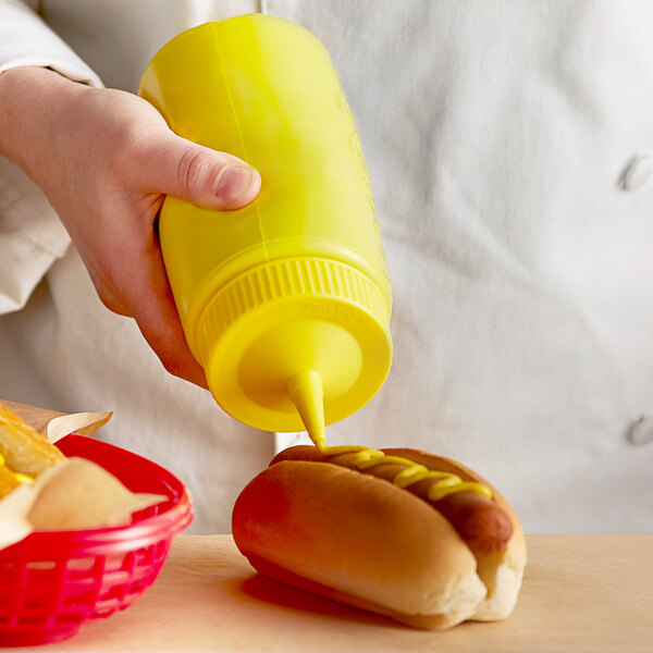 A hand holding a yellow plastic Vollrath squeeze bottle pouring mustard onto a hot dog.