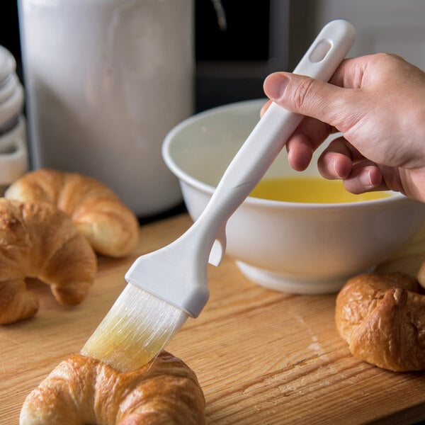 A hand holding a Carlisle Sparta Spectrum pastry brush over croissants.