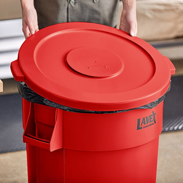 A person holding a red Lavex 44 gallon round commercial trash can lid.