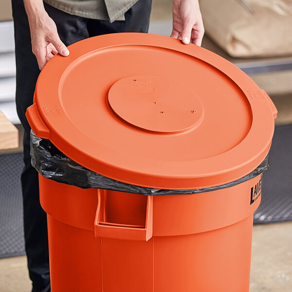 A person holding a Lavex orange plastic lid over a commercial trash can.