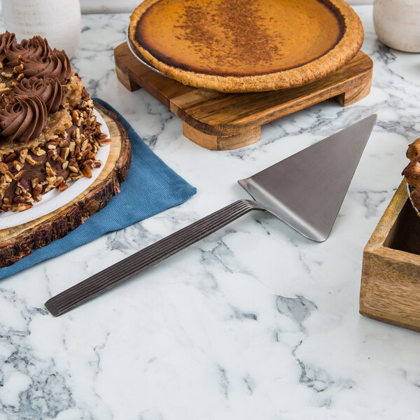 An American Metalcraft wavy stainless steel pie server cutting a cake on a marble surface.