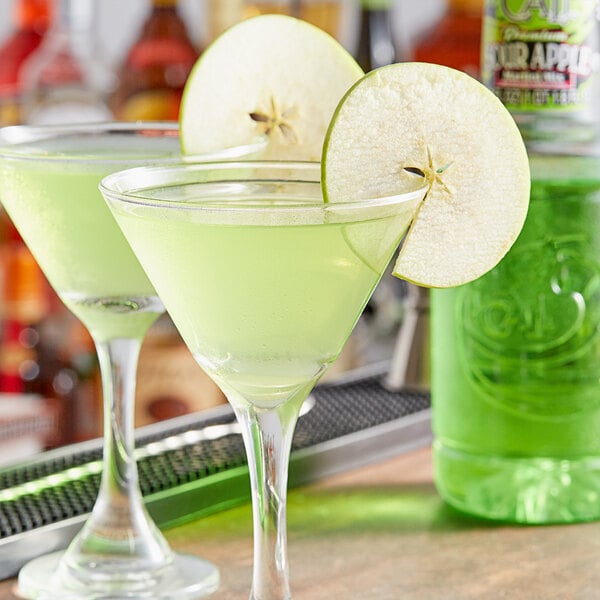 A table with two green martini glasses filled with green liquid and green apple slices with a bottle of vodka in the background.