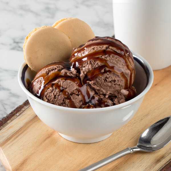 A Schonwald dark blue porcelain bowl filled with ice cream topped with chocolate syrup and cookies on a table with a spoon.