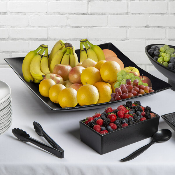 A black G.E.T. Enterprises Bugambilia square fruit bowl filled with bananas, apples, grapes, and a peach on a table with black tongs.