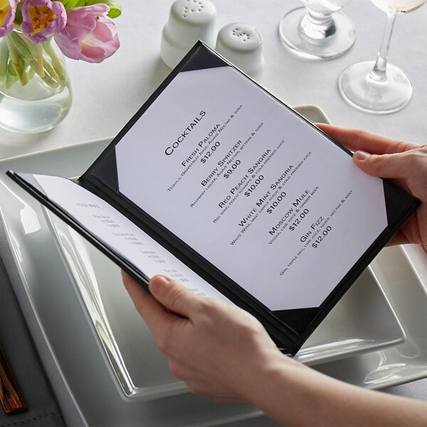 Hands holding a menu with a black cover on a table in a fine dining restaurant.