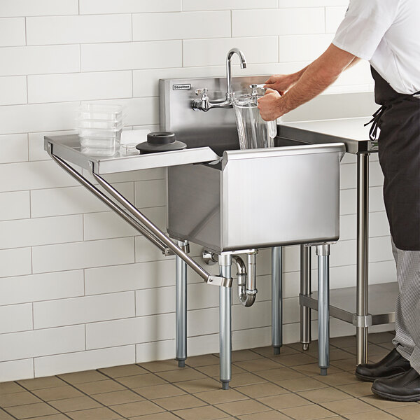 A man in a black apron washing clear plastic containers in a Steelton commercial utility sink.