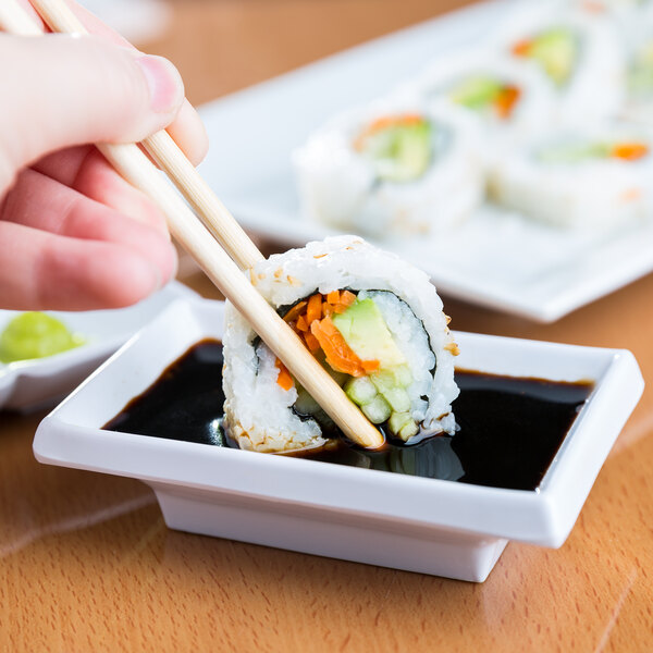 A person using chopsticks to dip a sushi roll into sauce in a white bowl.