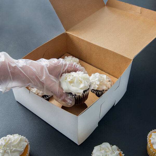 A hand in a plastic glove holding a white cupcake in a white box with a clear plastic window.