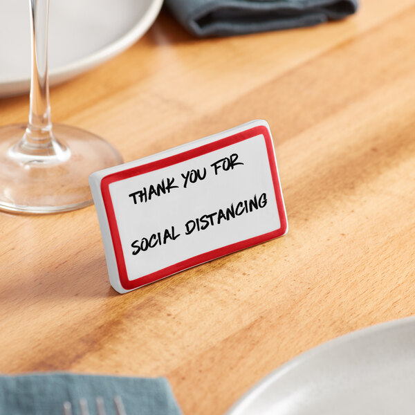 A red ceramic table tent sign with a border that reads "Thank you for social distancing" on a table.