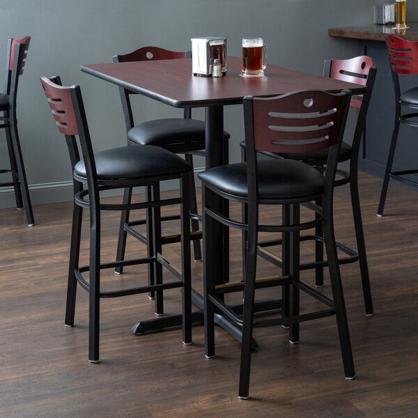 A Lancaster Table & Seating bar height cherry table with black chairs and padded seats with beer on it.