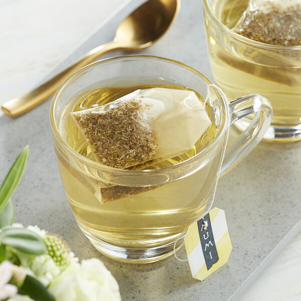 A glass mug of Numi Organic Chamomile Lemon tea with a tea bag in it next to a spoon.