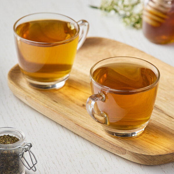 A wooden tray with two glass cups of Numi Moroccan Mint tea, a jar of honey, and a jar of herbs.