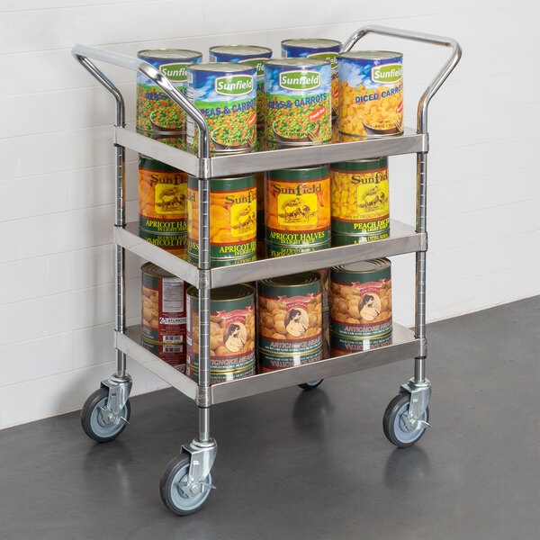 A Regency stainless steel utility cart full of canned peas, carrots, and soup.