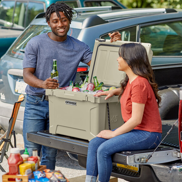 A man and woman sitting on the back of a truck with a CaterGator tan cooler.