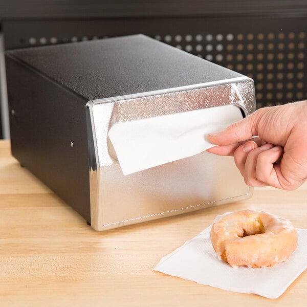 A hand using a Vollrath black countertop napkin dispenser to get a napkin for a donut.