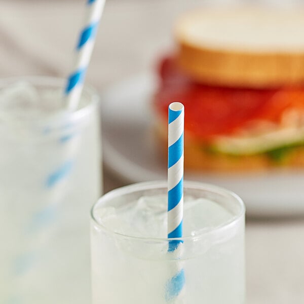 A close-up of a EcoChoice blue and white striped paper straw in a glass of ice with a sandwich on a plate.
