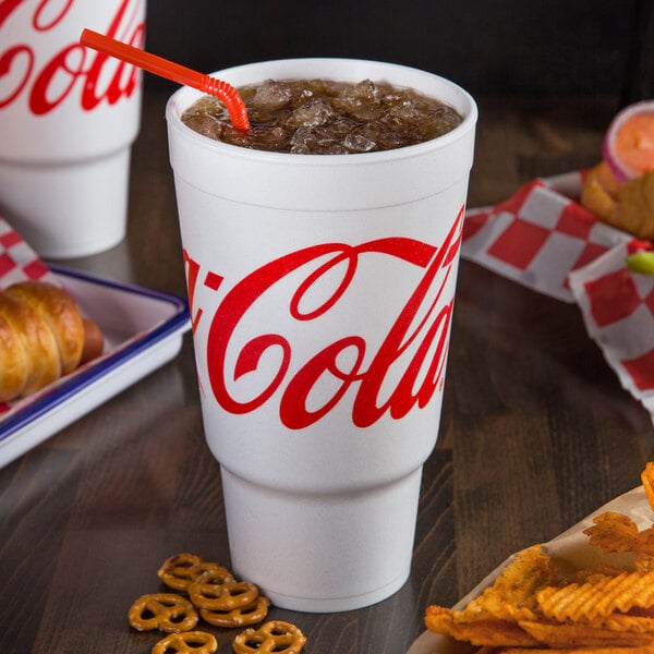 A Dart foam cup with Coke and a straw on a table with snacks.