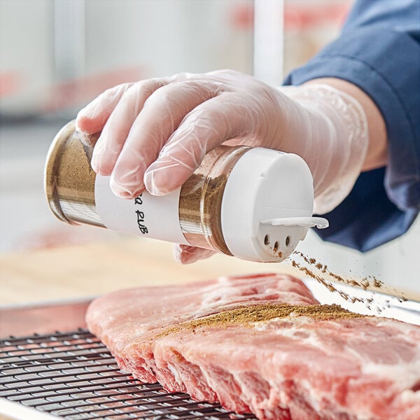 A gloved hand using a 53/485 round plastic spice container to season a piece of meat.
