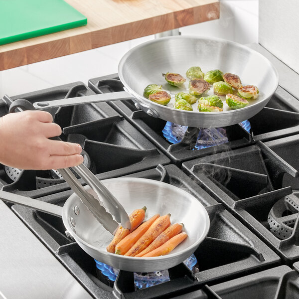 A person cooking carrots and brussels sprouts in a Choice aluminum fry pan on a stove with tongs.