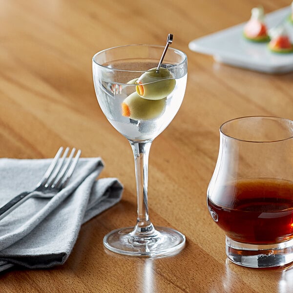 A Libbey Nick and Nora glass filled with brown liquid with olives on a table.