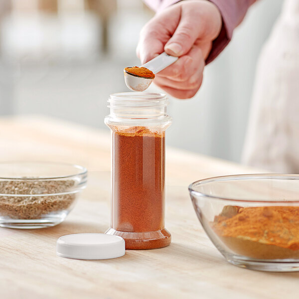 A person using a spoon to pour red chili powder into a round plastic spice container with a white lid.