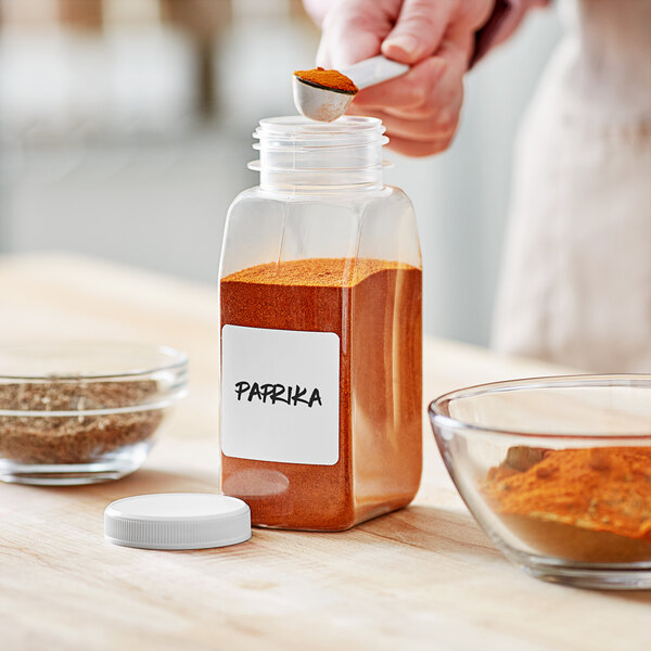 A person pouring paprika into a 53/485 rectangular plastic spice container with a white lid.